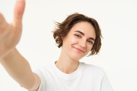 Lifestyle and people. Young woman smiling and taking selfie, posing for photo, holding camera with one hand, standing over white background.