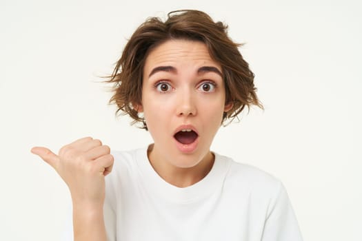 Close up portrait of brunette girl gasps and looks surprised, points left to inquire about promotion, stands over white background.