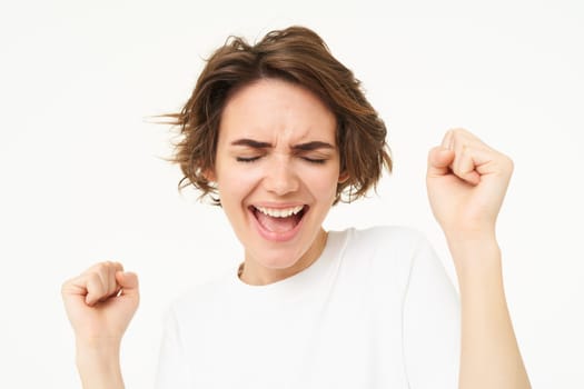 Image of happy, excited brunette woman, clenches her fists, dancing in excitement, feeling amused, standing over white background.