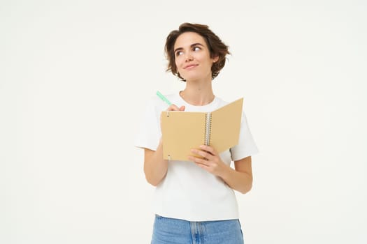 Portrait of thoughtful and creative woman writing down her thoughts in diary, doing homework, looking aside and thinking, posing over white background.