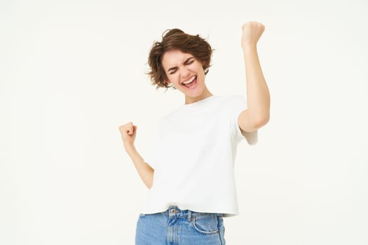 Cheerful brunette woman winning, triumphing, celebrating victory, achieves goal, stands over white background.