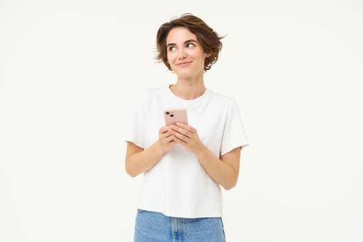 Portrait of brunette woman using smartphone, standing with mobile phone, texting, placing an order, browsing social media in telephone, standing over white background.