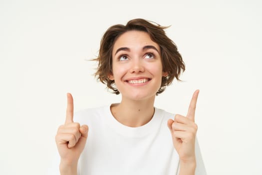 Portrait of happy, beautiful young woman pointing fingers up, showing advertisement or product on top of copy space, standing against white background.