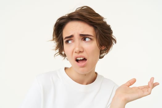 Portrait of disappointed young woman complaining, holding hand sideways, talking, expresses dislike and irritation, standing over white background.