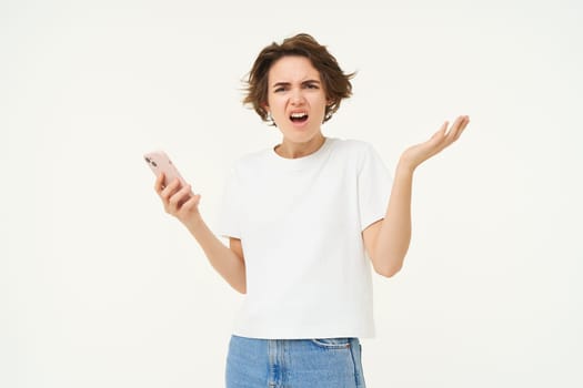 Portrait of confused and upset young woman, holding smartphone, shrugging shoulders and complaining at something online, standing over white background.