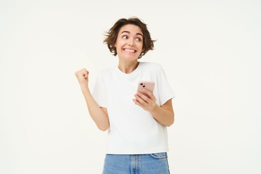 Portrait of brunette woman holding mobile phone, winning, triumphing after completing achievement on smartphone app, standing over white background.