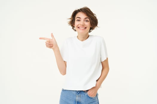 Image of cute woman with happy smile, pointing left, showing advertisement, standing over white background. Copy space