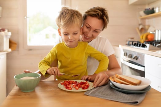 young caucasian blond mother and child son sitting in the morning in the kitchen and preparing breakfast with toast and strawberries.
