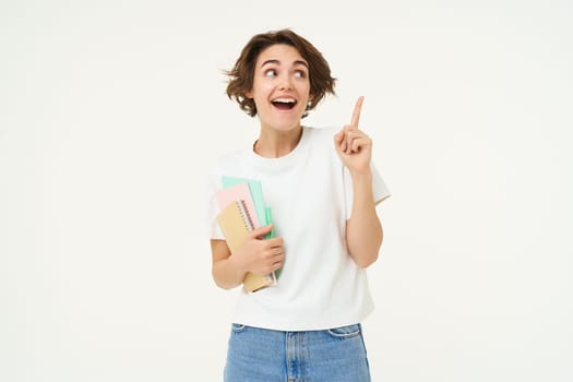 Portrait of brunette woman laughing, student with notebooks pointing at upper right corner, showing banner or advertisement, standing over white studio background.