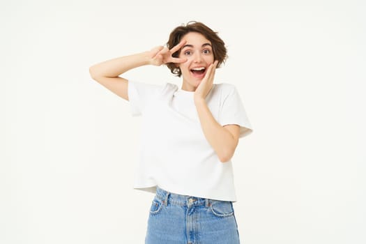 Portrait of happy and carefree woman, shows peace, v-sign gesture, having fun, posing for photo over white background.