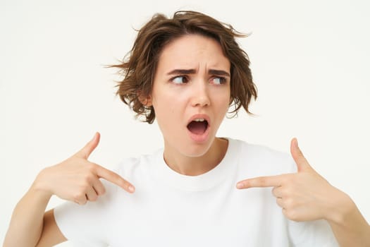 Insulted woman points at herself, being offended or hurt, looks shocked aside, stands over white studio background.