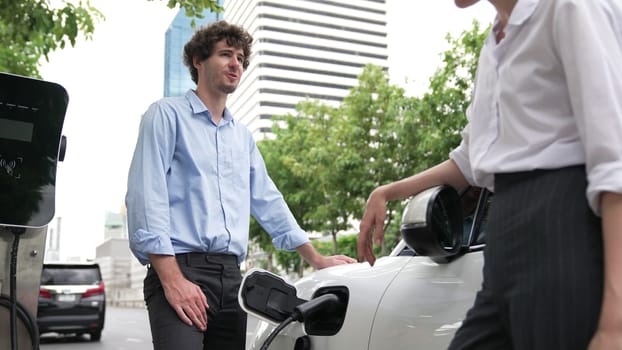 Progressive businessman and businesswoman install charger plug from charging station to electric car before driving around city center. Eco friendly rechargeable car powered by clean energy.