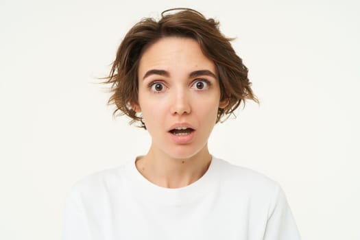 Close up shot of woman with shocked face, gasping and staring startled at camera, confused and surprised by something, standing over white background.