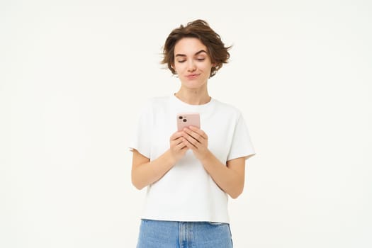 Portrait of brunette girl with smartphone, sending a message, using mobile phone application, smiling and looking happy, white studio background.