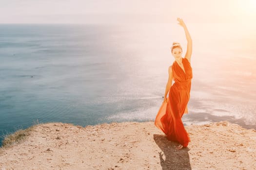 Side view a Young beautiful sensual woman in a red long dress posing on a rock high above the sea during sunrise. Girl on the nature on blue sky background. Fashion photo.