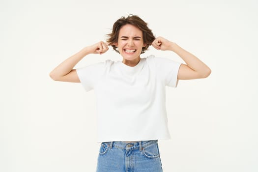 Portrait of upset woman shuts her ears, feeling discomfort from loud noise, standing over white background. Copy space