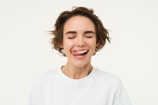 Close up of happy smiling woman, licks her white teeth, feels happy after dental treatment, isolated against studio background.