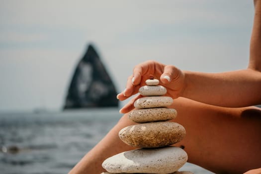 Woman bilds stones pyramid on the seashore on a sunny day on the blue sea background. Happy holidays. Pebble beach, calm sea, travel destination. Concept of happy vacation on the sea, meditation, spa