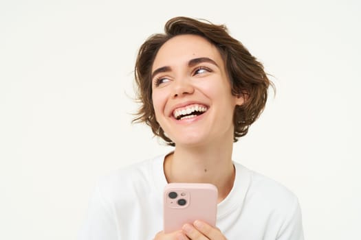 Portrait of young woman standing with pink mobile phone, using smartphone app, doing online shopping, paying for something using application, standing over white background.