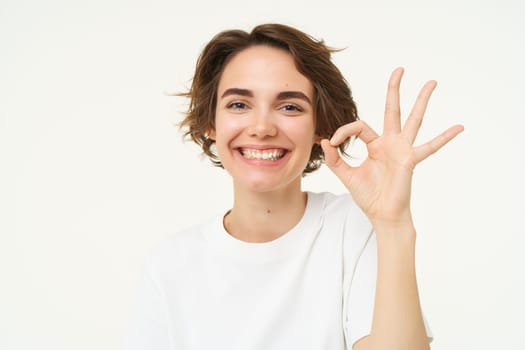 Close up portrait of satisfied, smiling young woman, shows okay, ok gesture, recommends something, gives positive feedback, says yes, white background.