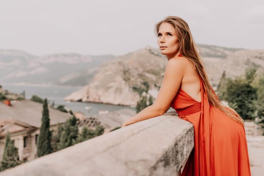 Side view a Young beautiful sensual woman in a red long dress posing on a volcanic rock high above the sea during sunset. Girl on the nature on overcast sky background. Fashion photo