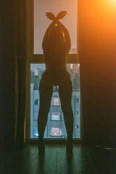 A full-length silhouette of young woman at open balcony window and dark blue night sky, with a cityscape in the background. Seduction and passion concept. Romantic moment in the bedroom. Night room