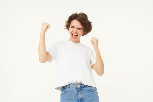 Excited brunette girl, student winning prize, celebrating victory, triumphing, standing in white t-shirt and jeans over white background.