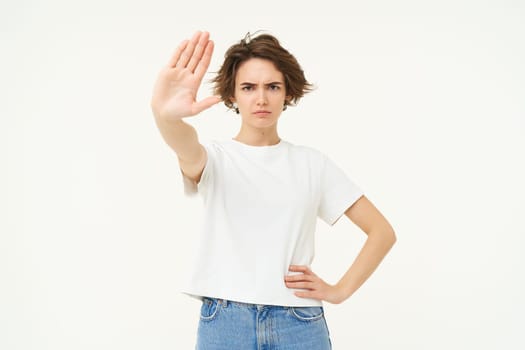 Image of serious young woman extends one hand, shows stop, prohibit gesture, forbid something, standing over white background.