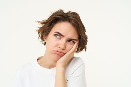 Portrait of young bored woman, leans head on hand and frowns, looks upset and grumpy, thinking, standing over white background.