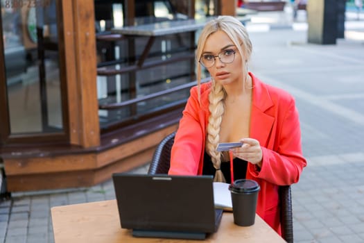 Beautiful woman sits at table in street cafe and works on tablet. Concept remote work, free work schedule. In a bright pink jacket and glasses