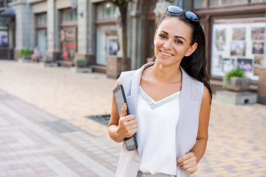 Confident young woman laughing and carrying notepad in hand while standing on the street, wearing stylish clothes