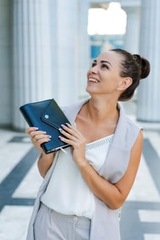 Confident young woman laughing and carrying notepad in hand while standing on the street, wearing stylish clothes