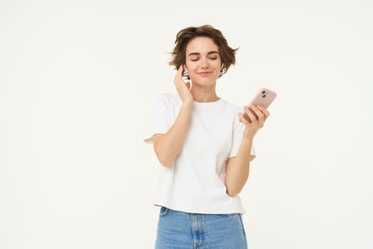 Portrait of happy brunette woman, holds smartphone, listens music in wireless headphones, stands over white studio background.