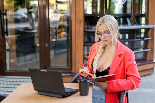 Beautiful woman sits at table in street cafe and works on tablet. Concept remote work, free work schedule. In a bright pink jacket and glasses