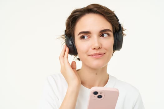 Portrait of candid young woman, listens music in wireless headphones, holding smartphone, watching videos on mobile phone, standing over white background.
