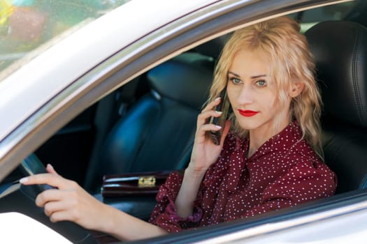 portrait of a beautiful blonde woman in a red dress sitting in a car with a phone