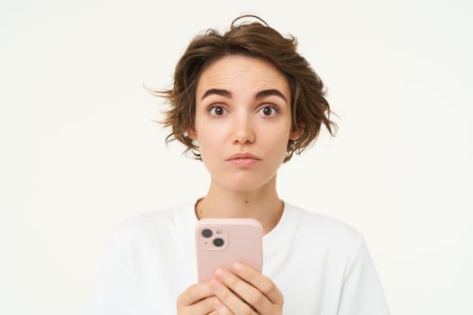 Portrait of confused girl standing with smartphone, raising her eyebrows in surprise, standing over white background.