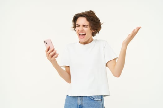 Portrait of woman singing and looking excited, holding mobile phone, triumphing, standing against white background.