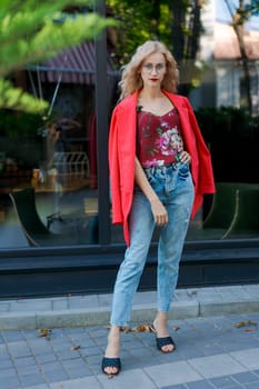 beautiful blonde woman in glasses dressed in red jacket and blue jeans posing on street in the city