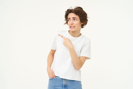 Portrait of woman looking with doubt, pointing left, staring at something with unsure, concerned face expression, standing over white studio background.