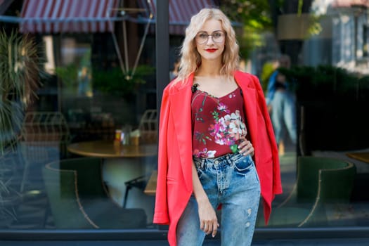 beautiful blonde woman in glasses dressed in red jacket and blue jeans posing on street in the city