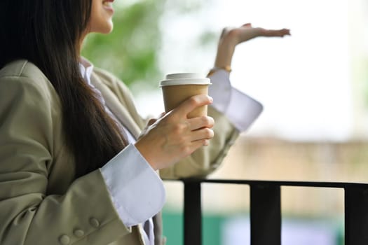 Relaxed businesswoman with paper cup of coffee catching raindrops with hand on office balcony.