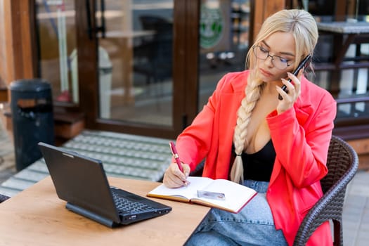 Beautiful woman sits at table in street cafe and works on tablet. Concept remote work, free work schedule. In a bright pink jacket and glasses