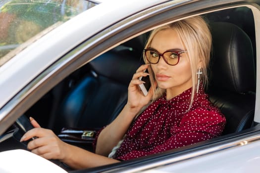 portrait of a beautiful blonde woman in a red dress sitting in a car with a phone