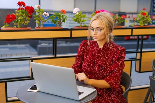 Beautiful woman works on laptop in coffee shop and using mobile phone in a red dress and glasses, business blonde online business