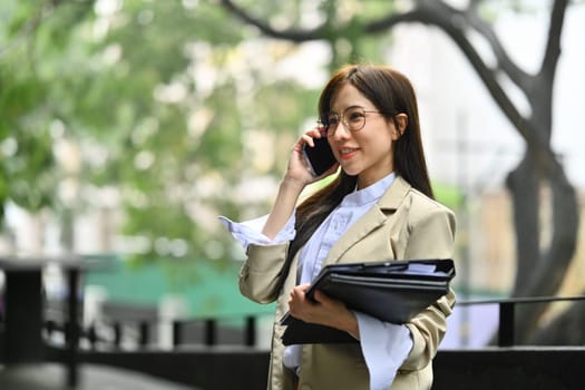 Beautiful millennial businesswoman having pleasant phone conversation while walking in the city.