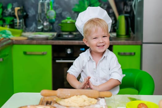happy little boy preparing dough in kitchen at table. there are dough products on table, dressed as chef