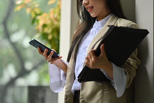 Pleasant businesswoman using smartphone chatting online with friends during break while standing outside office building.