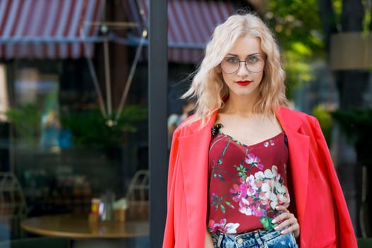 beautiful blonde woman in glasses dressed in red jacket and blue jeans posing on street in the city