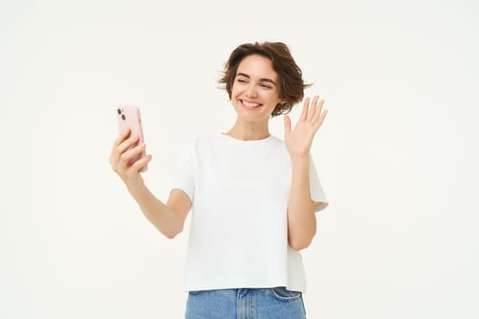 Portrait of woman video chats on smartphone, talking with someone on mobile phone app, waving hand at camera, standing over white background.